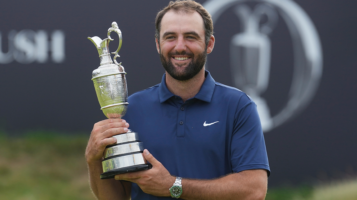 Scottie Scheffler smiling with the British Open Trophy in hands, turned to the left in front of the British Open banner.